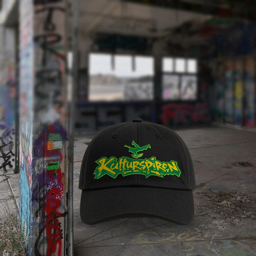 Svart kaps med Kulturspiren-graffitilogo på betonggulv foran graffitivegger